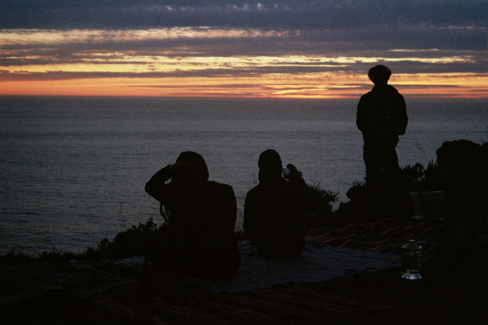 A silhouetted image of three people watching the sunset from a high bluff above the ocean